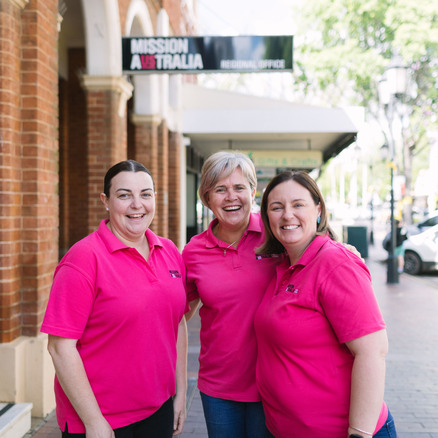 Three women wearing Mission Australia shirts smile while standing together on a city sidewalk in front of a brick building with a Mission Australia Regional Office sign.