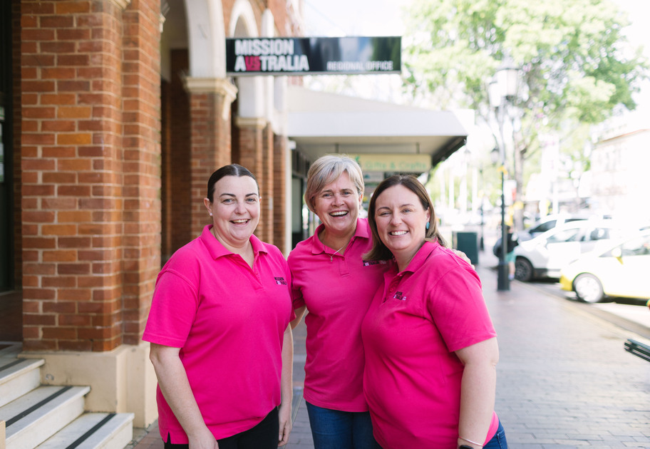 Three women wearing Mission Australia shirts smile while standing together on a city sidewalk in front of a brick building with a Mission Australia Regional Office sign.