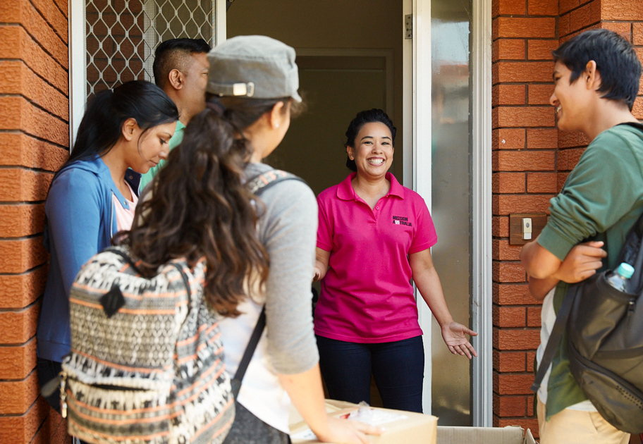 A Mission Australia case worker stands smiling at the door of a brick house, welcoming a group of young people with backpacks. A box sits in the foreground, suggesting they are delivering or receiving something.