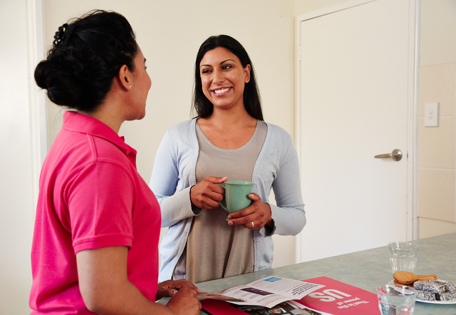 wo women standing in a kitchen and smiling at each other. One woman is holding a mug, while the other, a Mission Australia case worker is leaning on the counter next to an open magazine and a glass of water.