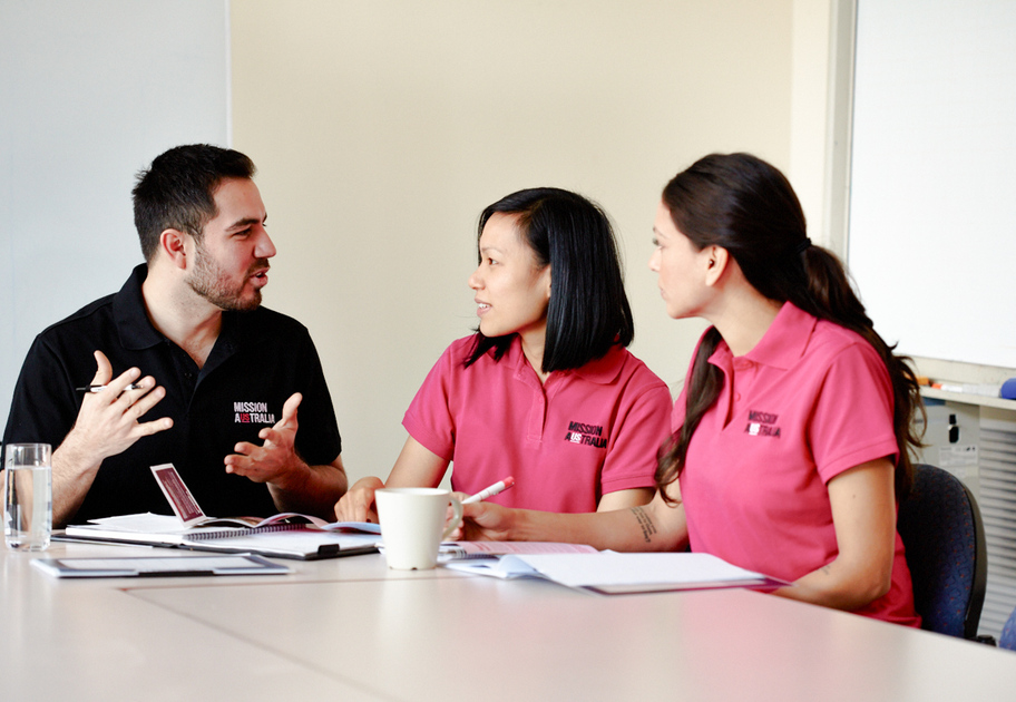 Mission Australia case workers sit at a table in discussion, with notebooks, a coffee cup, and a glass of water in front of them.