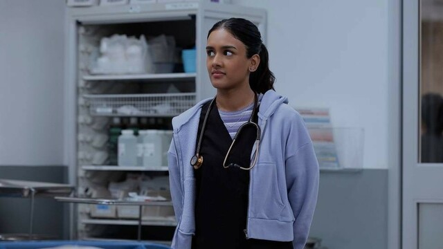 Victoria Javadi stands in a hospital room wearing scrubs and a hoodie, listening attentively.