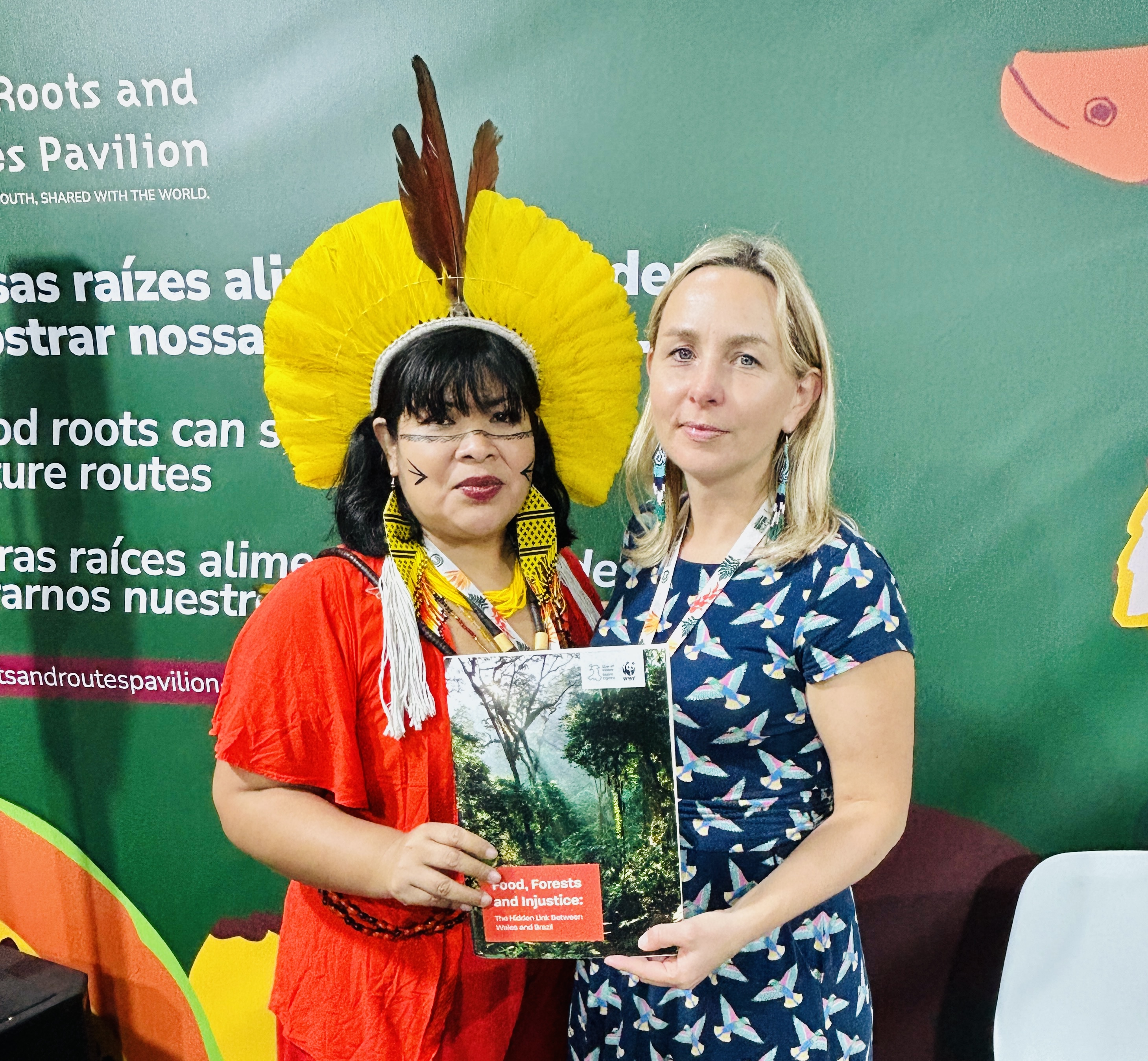 Two women, one in traditional Indigenous attire and one in a blue patterned dress, hold the 