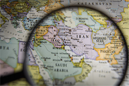 A close-up photo of a political world map with a magnifying glass centered over the Middle East. The lens focuses on Iran, Iraq, and the Persian Gulf, making city names like Tehran, Baghdad, and Kuwait City clearly legible. The surrounding areas, including parts of Pakistan, Afghanistan, and Saudi Arabia, are visible but slightly blurred outside the magnification.