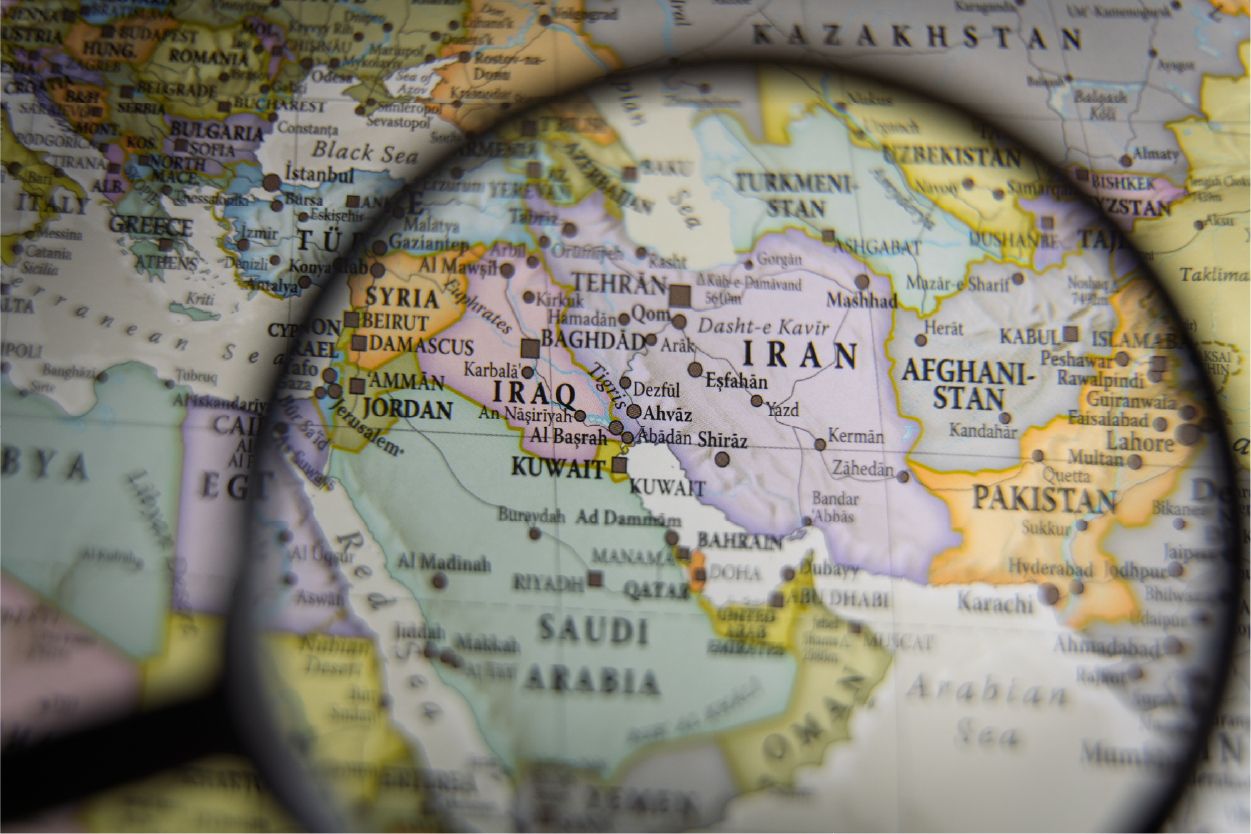 A close-up photo of a political world map with a magnifying glass centered over the Middle East. The lens focuses on Iran, Iraq, and the Persian Gulf, making city names like Tehran, Baghdad, and Kuwait City clearly legible. The surrounding areas, including parts of Pakistan, Afghanistan, and Saudi Arabia, are visible but slightly blurred outside the magnification.