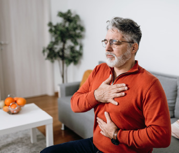 Starejši moški v rdečem puloverju sedi na kavču in zdi se, da občuti znake srčnega infarkta – bolečino v prsih in slabost.