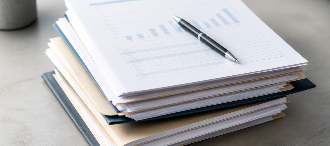 Stacks of folders and paperwork with a page of a bar graph on top and a pen on top of that page.