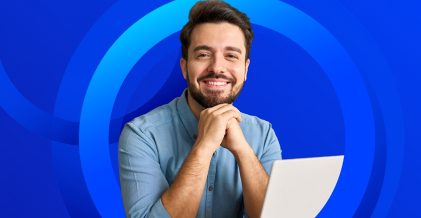 A smiling, bearded man in a light blue shirt is looking directly at the camera with his hands clasped under his chin, sitting in front of a white laptop against a bright blue background with concentric rings.