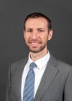 Headshot of Ted Mulrane, who smiles while wearing a light grey suit and a blue tie