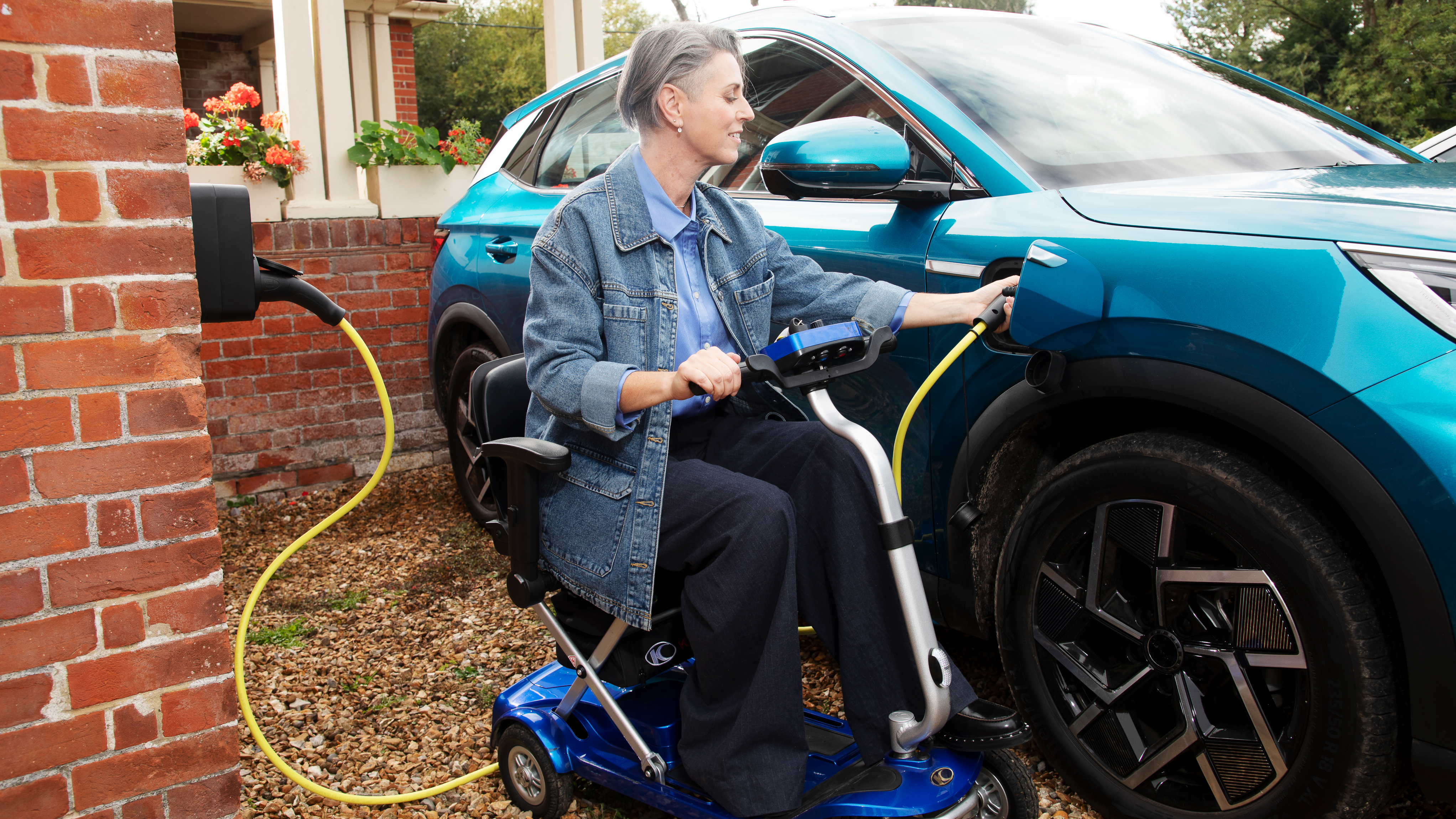 A person using a mobility scooter plugs a charging cable into a blue electric car outside a home.