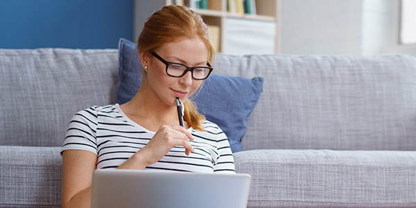 A young white woman sits holding a pen and looking thoughtfully at a laptop screen