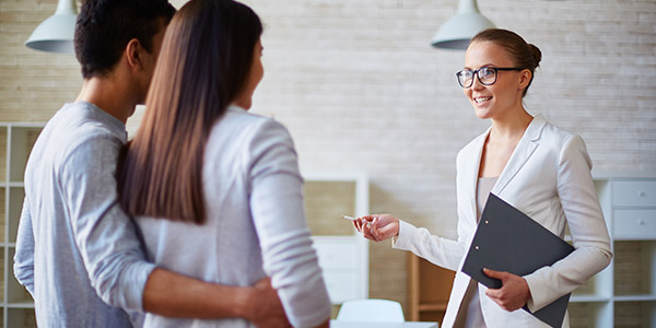 White woman in a business suite holds a clipboard and speaks to a couple in a home