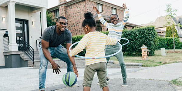 A Black man plays with his two kids in front of their home