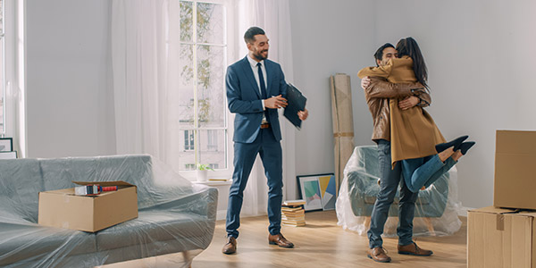 A smiling man pics a woman up in a hug inside of a home while a professional stands beside them smiling 