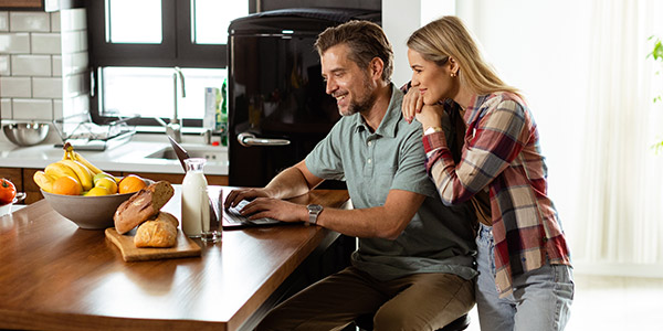 A man and a woman smile while they look at the laptop that is sitting on their kitchen counter