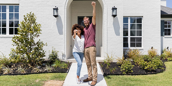 a Black woman and a white man stand together smiling and holding house keys in front of their new home