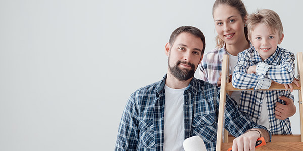 A white man, woman, and child all stand smiling on a ladder together. The man is holding a paint roller.