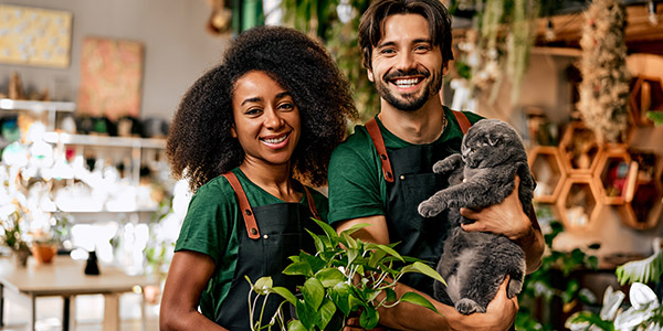 A Black woman and a white man wearing aprons for work standing smiling together in a shop. The man is holding a cat. 