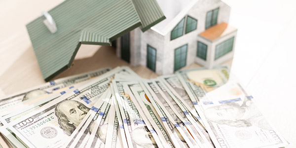 A model of a house sits on a table with a stack of hundred dollar bills