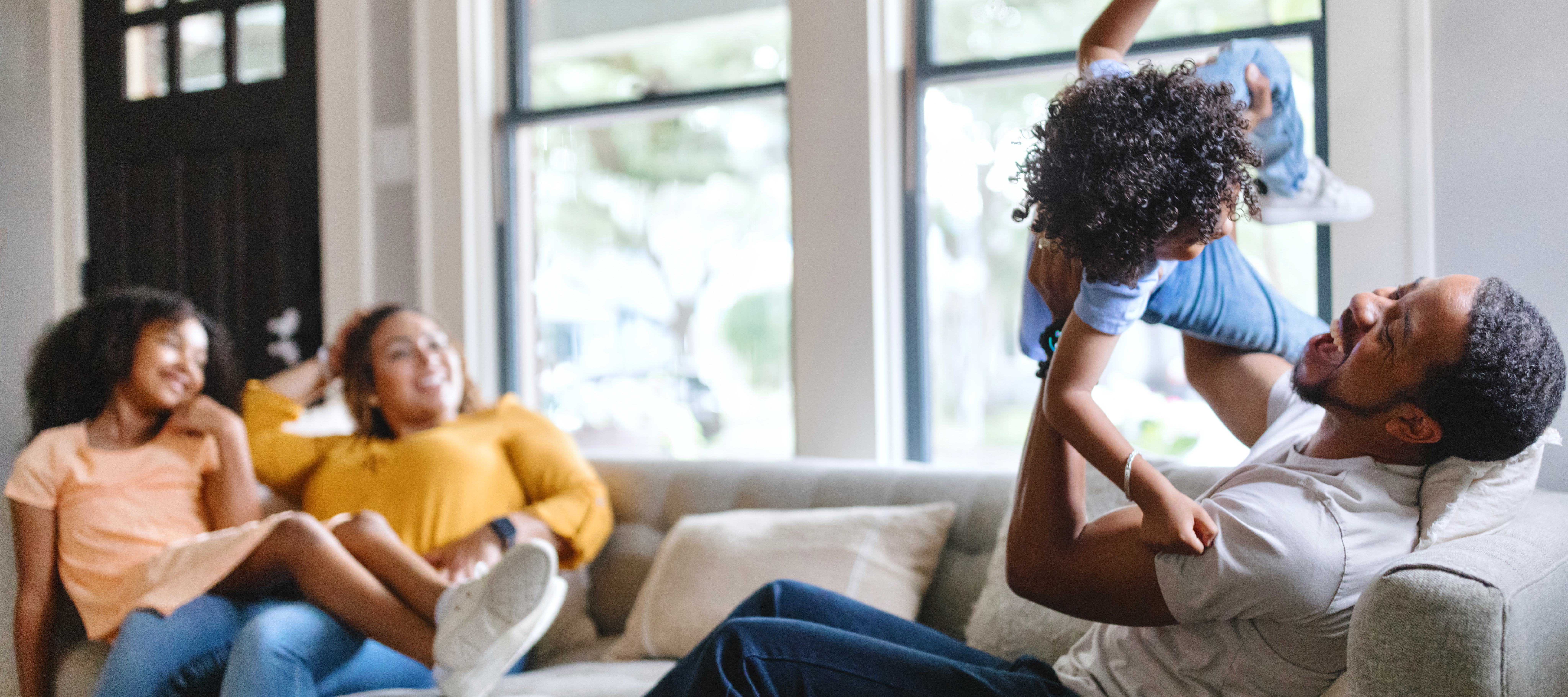A Black man plays with his daughter in their living room while two other Black women sit on the couch smiling 