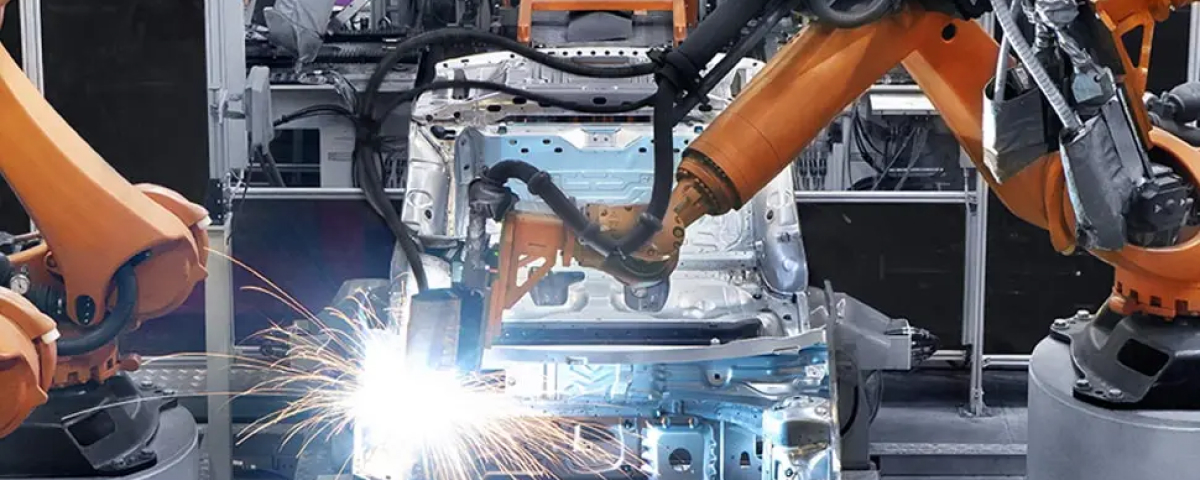 An image of an automated factory line with three large, orange industrial robotic arms assembling a car chassis. Bright sparks fly from a welding point in the center of the frame.