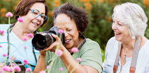 Three smiling women photographing pink flowers in a lush garden while holding professional cameras.