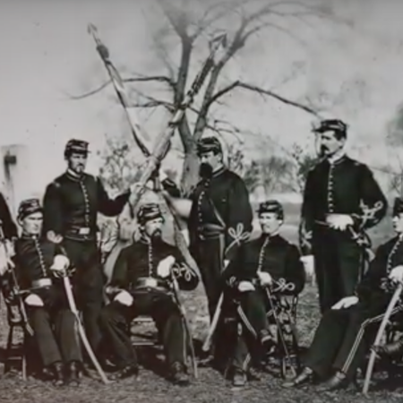 Civil war era military members stand and sit in chairs while posing for a black and white, grainy photo.
