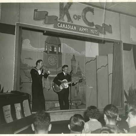 Priest singing with guitarist in a WW2 army hut operated by Canadian Knights.