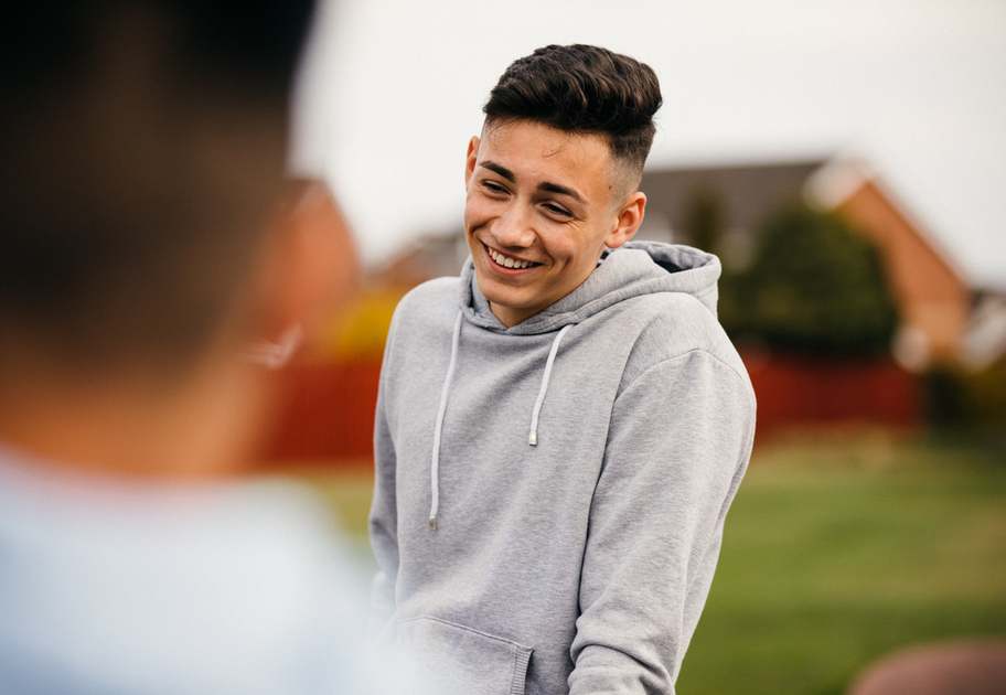 A teenage boy wearing a gray hoodie smiles while talking to someone outdoors, with houses and greenery visible in the blurred background.