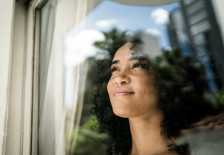 A woman with curly hair looks thoughtfully out a window, with reflections of clouds and trees on the glass, blending her face with the outdoor scenery.